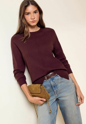 Jeune femme portant un pull bordeaux et un jean bleu clair, tenant un petit sac à main en suède marron avec une bandoulière en chaîne dorée.