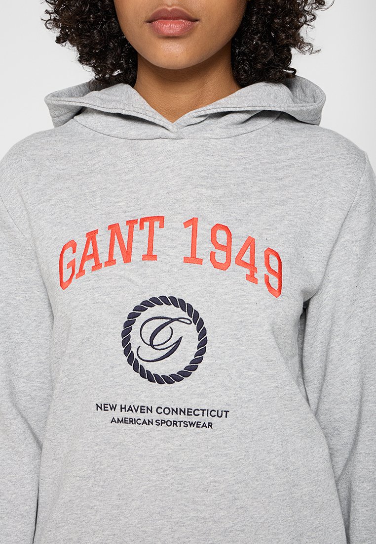 Grauer Kapuzenpullover mit „GANT 1949“ in oranger Schrift, einem marineblauen kreisförmigen Logo mit verwobenem Seil und „NEW HAVEN CONNECTICUT“ in schwarzem Text.