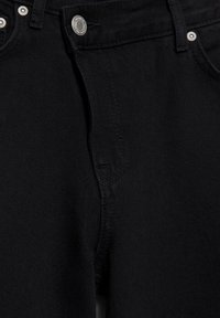 Gros plan sur un jean en denim noir montrant le bouton, la zone de la fermeture éclair, les passants de ceinture et les poches avant avec des rivets métalliques.