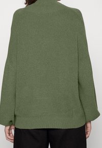 Zielony sweter z dzianiny z wysokim kołnierzem, szerokimi rękawami i prążkowaną fakturą. Projekt cechuje się luźnym krojem oraz lekko wydłużonym tyłem.