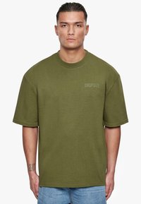 Grünes Baumwoll-T-Shirt mit fallenden Schultern, einem runden Halsausschnitt und "DROPSIZE" in einem dezenten Farbton auf der Brust bedruckt. Kurze Ärmel.