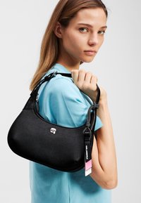 Schwarze Lederhandtasche mit runder Form, strukturiertem Oberflächenmaterial, silberner Hardware und einem abnehmbaren Riemen, der mit einem pinkfarbenen und weißen Anhänger versehen ist.
