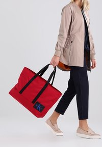 Borsa tote in tela rossa con manici neri e logo blu. La borsa presenta un design spazioso e un fondo piatto per garantire stabilità.
