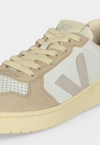 Χαμηλού τύπου μπεζ και λευκό αθλητικό παπούτσι με σουέντ λεπτομέρειες, κρεμ κορδόνια, διάτρητη περιοχή μύτης και μπεζ λογότυπο "V" στην πλαϊνή πλευρά.