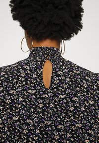 Blusa preta com padrão floral, apresentando um colarinho alto franzido, detalhe de abertura nas costas e pequenos padrões de flores multicoloridas por todo o tecido.