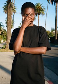 Rövid hajú személy fekete pólóban, aki részben az arcát takarja a kezével, napsütéses járdán áll, pálmafák a háttérben.