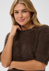 Jeune femme aux cheveux blonds portant un pull court à manches courtes marron foncé en matière pelucheuse, posant la main près du visage, souriant doucement sur un fond uni.