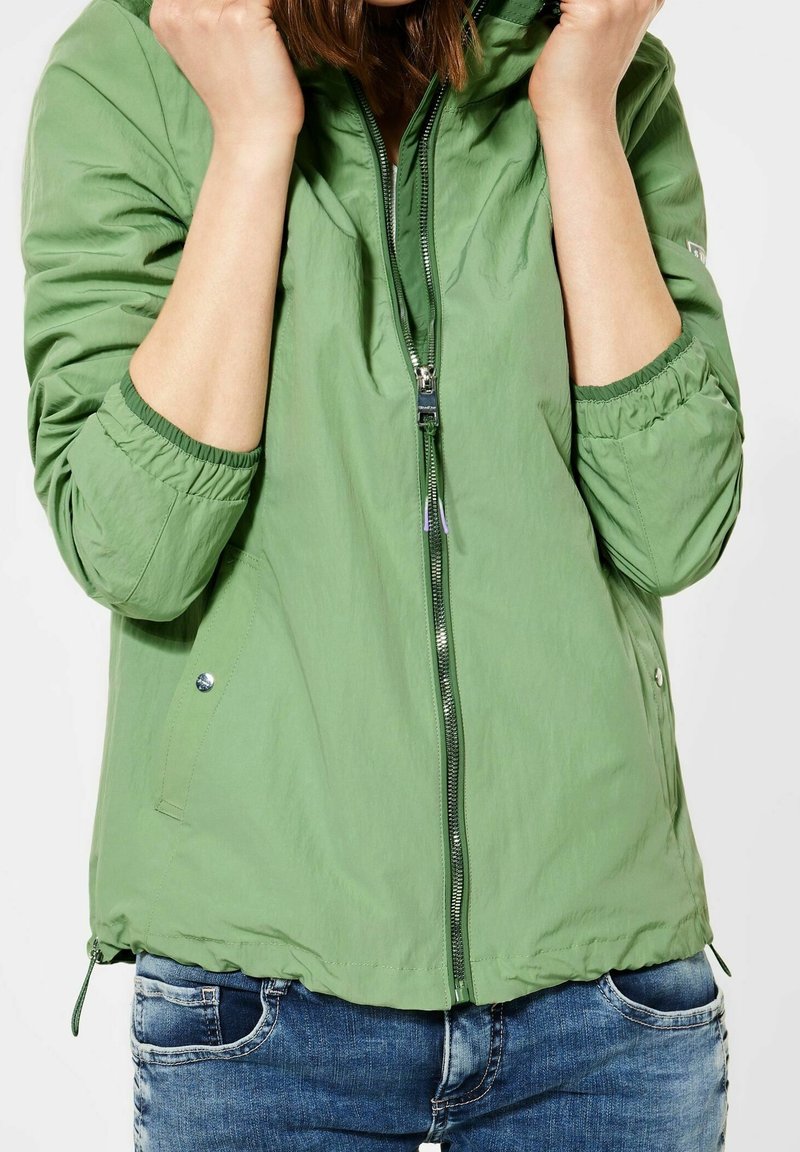 Grön lättvikts zip-jacka med elastiska ärmslut, två sidfickor och drar ihop nederkanten. Tygets textur verkar slät och något glänsande.