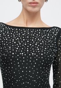 Κοντινό πλάνο ενός ατόμου που φοράει μαύρο τοπ διακοσμημένο με μικρά και μεγάλα αντανακλαστικά ασημένια καρφιά, και ασημένια σκουλαρίκια κρίκους.