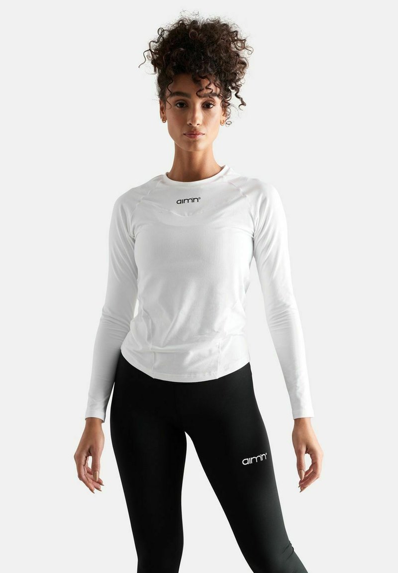 Hvid langærmet atletisk top med rund halsudskæring og subtile syninger, parret med sorte leggings med et logo. Glat tekstur.