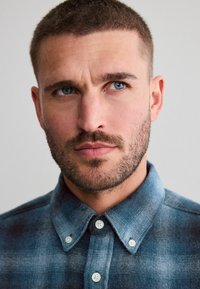 Cămașă din flanel în carouri albastre cu nasturi albi, având un guler de tip spread și o textură moale, fotografiată îndeaproape pe partea superioară a corpului unui model.