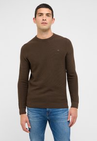 Pull en maille marron avec un col rond, des poignets et un ourlet côtelés. Présente un logo discret sur la poitrine, associé à un jean bleu clair.