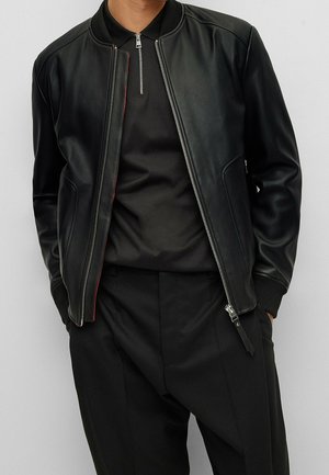 Mann trägt schwarze Leder-Bomberjacke über schwarzem Reißverschlusshemd und schwarzen Hosen, Hände in den Taschen, steht vor einfarbigem grauem Hintergrund.