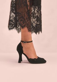 Sorte ruskinds-højhælede pumps med bølget design, fremhævet af en ankelrem og placeret mod en blød lyserød baggrund.