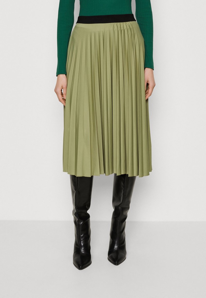 Falda midi plisada en tejido verde oliva con una cinturilla elástica negra, combinada con botas negras hasta la rodilla.