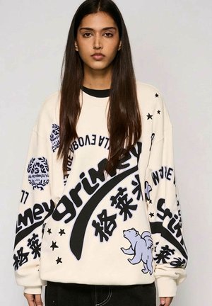 Młoda kobieta z długimi brązowymi włosami ubrana w biały, oversize'owy sweter z czarnym napisem, gwiazdami oraz małą grafiką kreskówkowego misia.