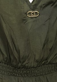 Abito in tessuto verde oliva con scollo a V, vita elasticizzata e una fibbia decorativa in argento sul busto. Tessuto liscio e materiale leggero.