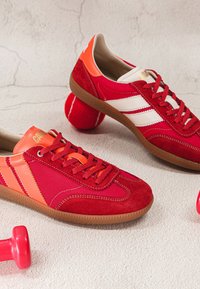 Paire de baskets rétro rouges et orange à côté d'un petit haltère rouge et d'une balle de tennis sur un sol clair texturé.