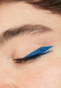 Blauwe glitter eyeliner aangebracht in een scherpe vleugelvorm op het gesloten ooglid, contrasterend met de natuurlijke huidtint en donkere wimpers.