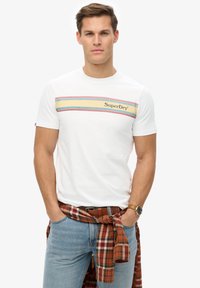 Camiseta blanca con un patrón de rayas horizontales multicolores y logo de "SuperDry". Llevada con jeans azul claro y una camisa de cuadros rojos anudada a la cintura.