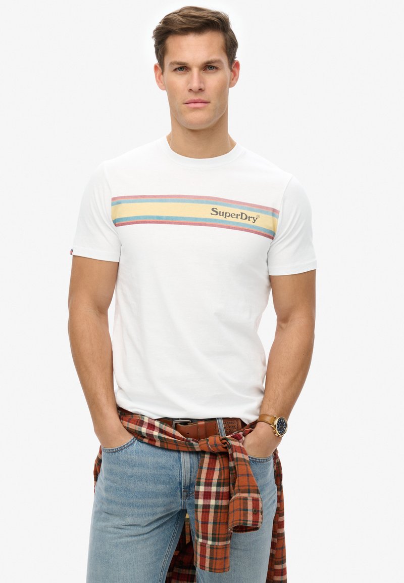 Camiseta blanca con un patrón de rayas horizontales multicolores y logo de "SuperDry". Llevada con jeans azul claro y una camisa de cuadros rojos anudada a la cintura.
