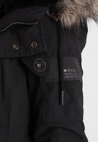 Sorte isolerede jakke med en pelsforet hætte, lynlås foran, trykknap og knaplukning, justerbar snor og et logo-patch på ærmet.