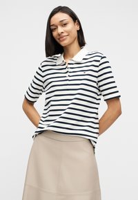 Γυναίκα με ίσια μαύρα μαλλιά που φοράει λευκό και ναυτικό μπλε ριγέ πόλο μπλουζάκι και μπεζ φούστα, στέκεται με τα χέρια πίσω από την πλάτη.