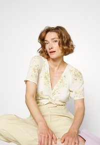 Blusa floral de mangas curtas com decote em V profundo e frente torcida, apresentando padrões de flores amarelas e verdes sobre um fundo claro.