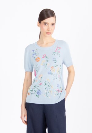 Kobieta o ciemnych włosach w jasnoniebieskiej bluzce z krótkim rękawem z wielokolorową kwiatową haftowaną aplikacją, w połączeniu z ciemnoniebieskimi spodniami, stojąca z jedną ręką w kieszeni.