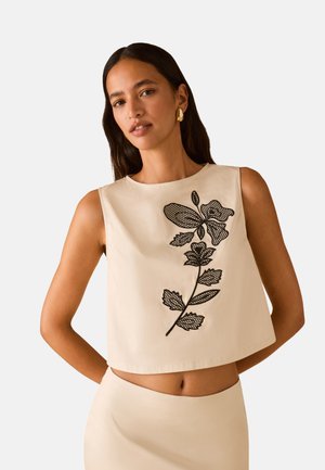 Vrouw met lang donker haar draagt een mouwloos beige cropped top met zwarte gebloemde borduur en een bijpassende hoog getailleerde rok, poseert met handen achter de rug.