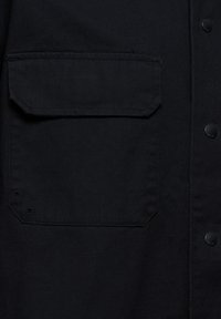 Nahaufnahme eines schwarzen Stoffs mit einer großen quadratischen Tasche, die eine Klappe und zwei Metallnieten auf der unteren linken Seite aufweist.