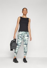 Top in maglia nera a coste, leggings marmorizzati verdi e neri, sneakers nere, mentre tiene una borsa grigio scuro. Abbigliamento sportivo semplice e aderente.