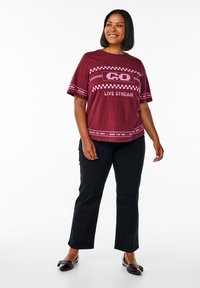 Bordeaux rood t-shirt met korte mouwen, voorzien van roze grafische tekst en ruiten, gecombineerd met zwarte broek. Het t-shirt heeft een relaxed fit en een ronde hals.