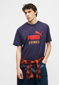 Νέος άνδρας που φοράει μπλε σκούρο μπλουζάκι Puma King με κόκκινο λογότυπο, κόκκινο-μαύρο πουκάμισο σε καρό δεμένο γύρω από τη μέση, και μπλε τζιν, κοιτάζοντας προς τα πλάγια.