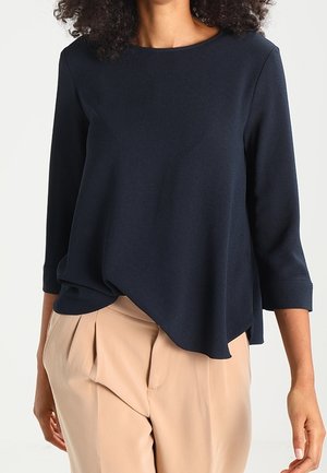 Vrouw draagt een marineblauwe top met asymmetrische zoom en lange mouwen, gecombineerd met beige plissébroek, zichtbaar van nek tot heupen.