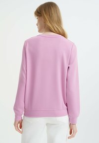 Felpa rosa con scollo rotondo e maniche lunghe. Morbida e liscia al tatto, presenta un orlo e polsini a costine. Indossata con pantaloni bianchi.