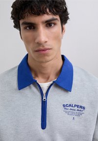 Jonge man met krullend donker haar, gekleed in een grijze ritshemd met blauwe kraag en 'Scalpers' tekst op de borst, op een effen lichte achtergrond.