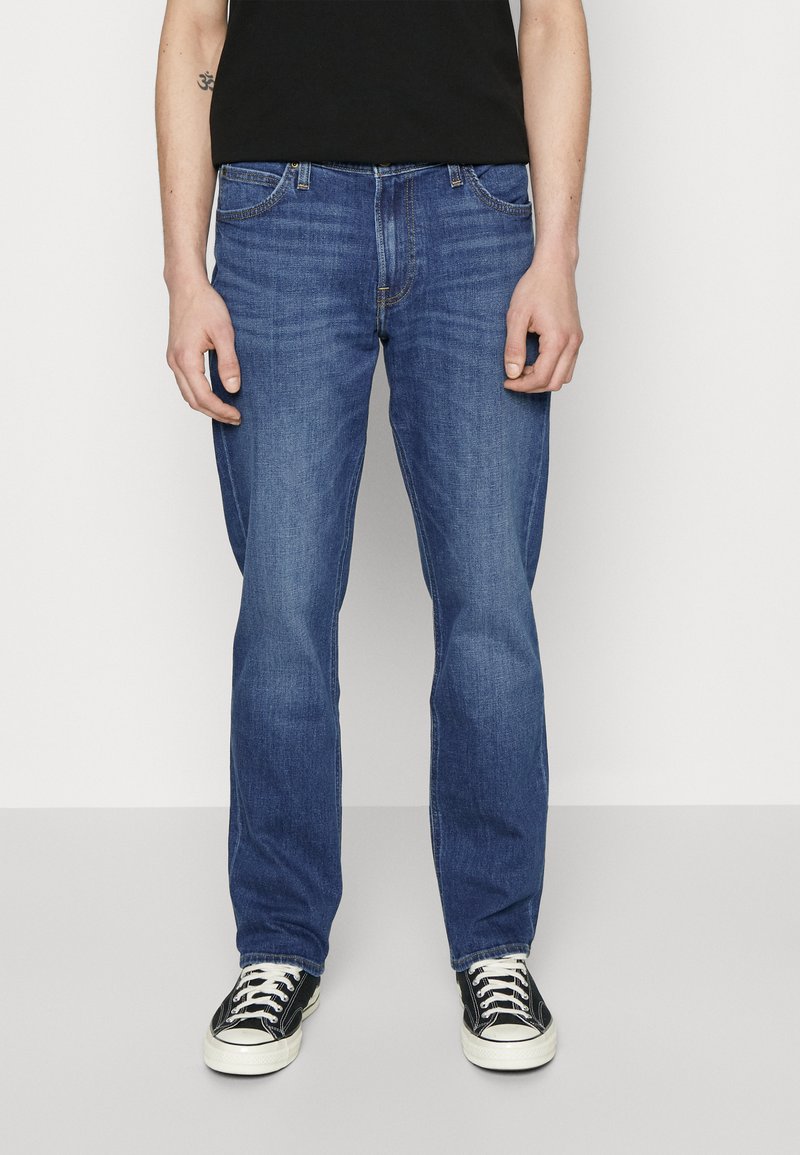 Jeans in denim blu con una vestibilità a gamba dritta, cinque tasche e sfumature delicate. Indossati con sneakers nere con lacci bianchi.