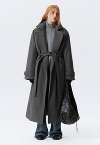 Lungo cappotto grigio con vita cintrata, ampi rever e tasche laterali, indossato sopra un maglione grigio. Tiene una borsa nera texturizzata. Jeans blu.
