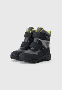 Bottes noires et grises avec une finition texturée, équipées de sangles Velcro et de accents jaunes vifs. Semelle robuste pour une adhérence sur différentes surfaces.