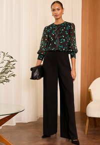 Pantaloni neri a gamba larga abbinati a una blusa nera trasparente con pois verdi e bianchi, e un dettaglio a fiocco al collo. Tiene una clutch nera.