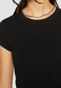 Svart bomullströja med korta ärmar och rund halsringning. Har en slät struktur och bärs med ett metallic kedjehalsband.