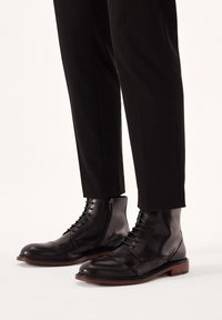 Botas de tobillo de cuero negro con un acabado suave, con cordones y una cremallera lateral, combinadas con pantalones negros ajustados.