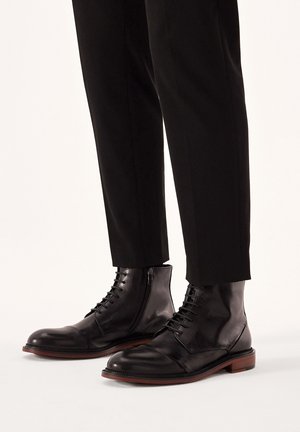 Botas de tobillo de cuero negro con un acabado suave, con cordones y una cremallera lateral, combinadas con pantalones negros ajustados.