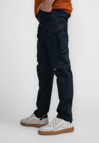 Marine cargobroek met meerdere zakken, gemaakt van duurzaam stof. Heeft een relaxte pasvorm en subtiele stikdetails. Gepaard met witte sneakers.