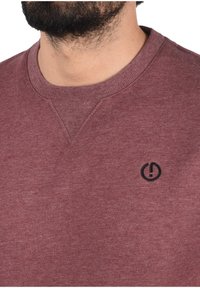 Maroon sweatshirt with a ribbed crew neckline and a small black logo on the chest. Soft texture with a casual design.