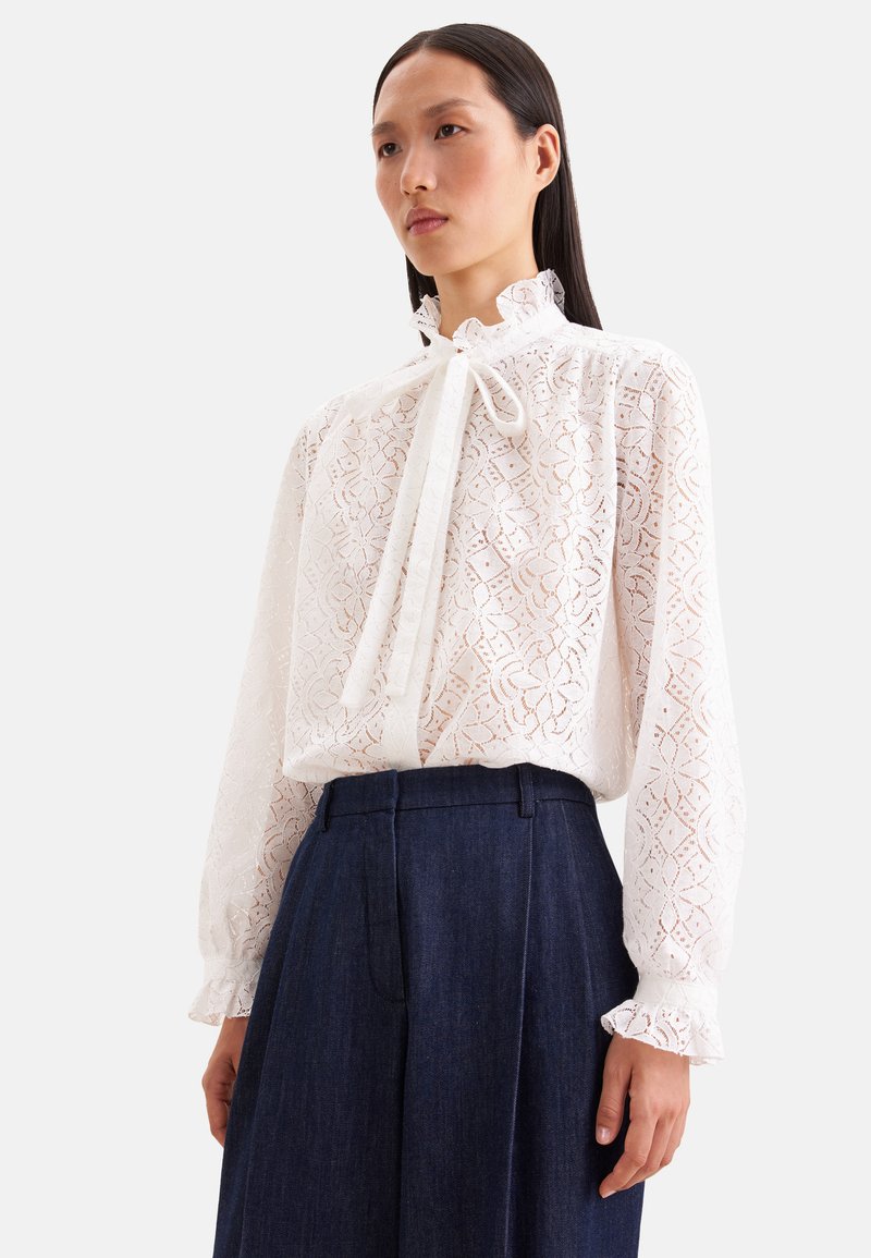 Femme portant un chemisier en dentelle blanche avec un nœud au col et des manches longues, associé à un pantalon bleu foncé taille haute.