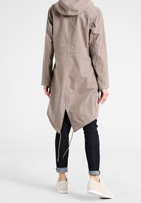 Impermeabile beige con cappuccio e orlo asimmetrico, caratterizzato da una texture liscia, maniche lunghe e vestibilità ampia, abbinato a jeans scuri e sneakers chiare.