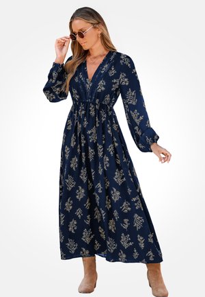 Abito lungo blu navy con maniche lunghe, scollatura a V e stampa floreale dorata; vita arricciata e gonna fluida; calzature in tonalità neutra.