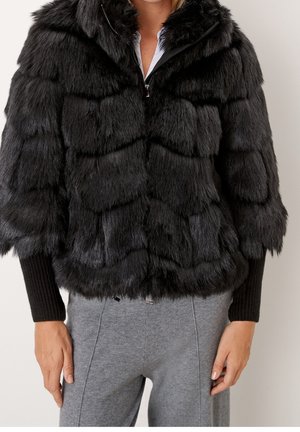 Frau trägt eine schwarze Jacke aus Kunstfell mit gerippten Bündchen und graue Hose, stehend vor einem schlichten Hintergrund.